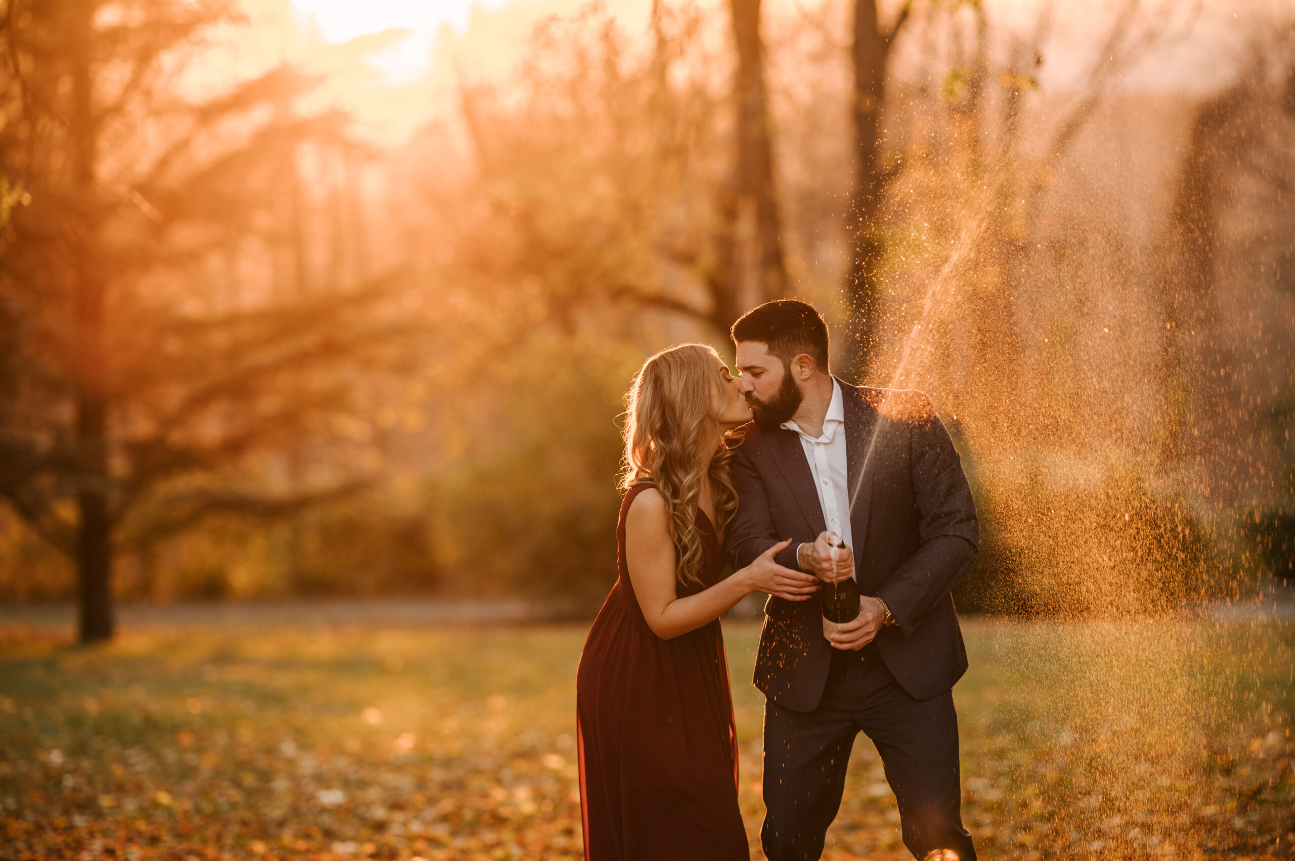Couple pops champagne at sunset during their engagement session at the New Jersey Botanical Gardens in Ringwood NJ. The Castle at Skylands Manor during golden hour