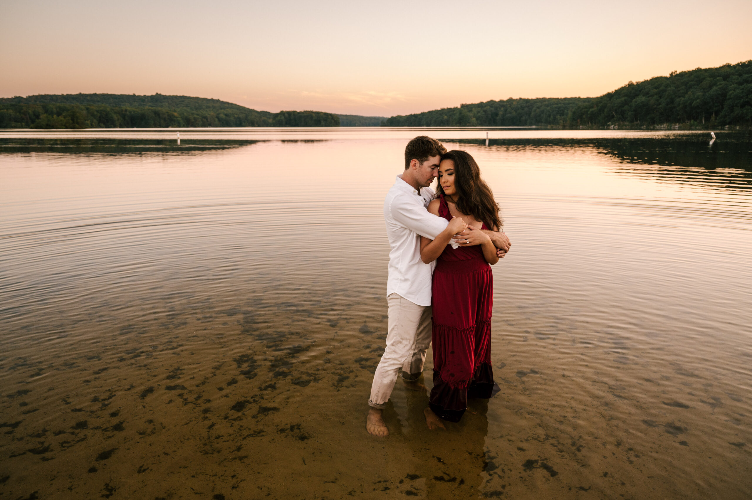 Couple standing in lake embracing, bride-to-be in red dress. Golden hour sunset at Wawayanda State Park in Hewitt, New Jersey
