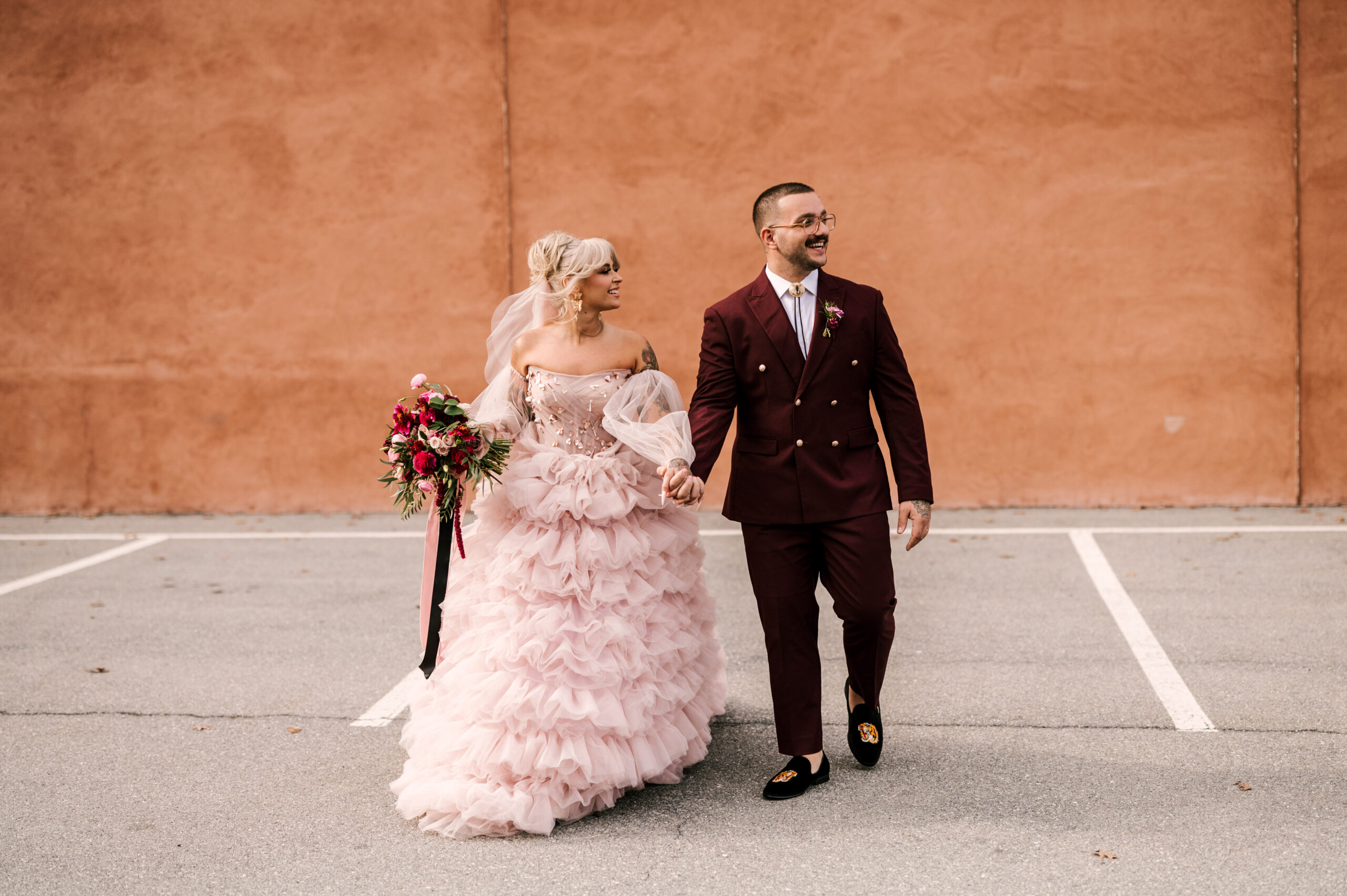 Alternative punk Bride and Groom walking together at their Cork Factory Hotel wedding in Lancaster Pennsylvania. Bride is in a pink punk rock barbie doll style dress with vibrant flowers. Groom is in burgundy suit. Industrial October wedding