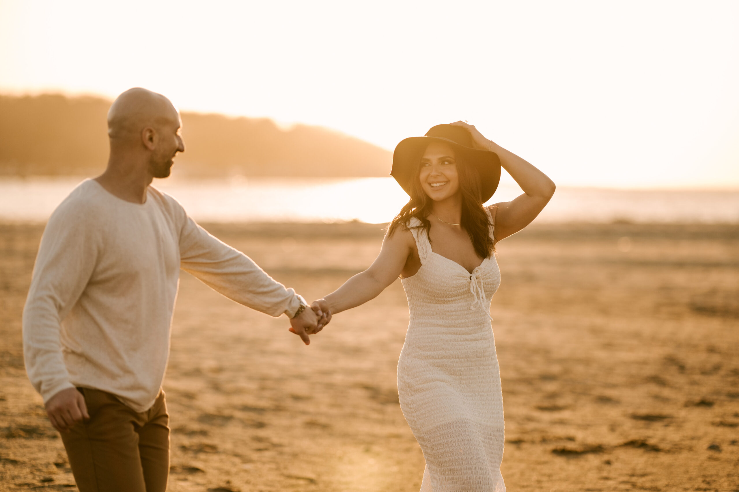couple at their engagement session holding hands and walking away from the water at sunset in sandy hook bay