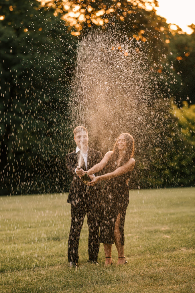 The newly-engaged couple pop open a champagne bottle with smiles on their faces in a field at Skylands Manor in Ringwood, NJ.