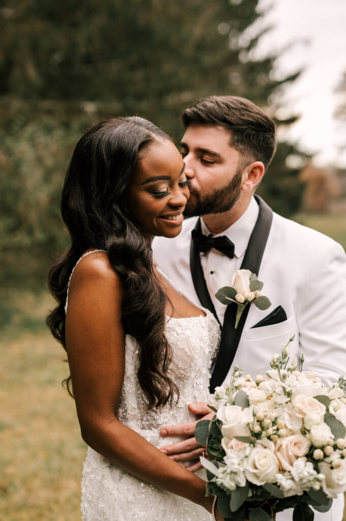 The groom kissing the bride's cheek as she smiles and looks down at Perona Farms Refinery.