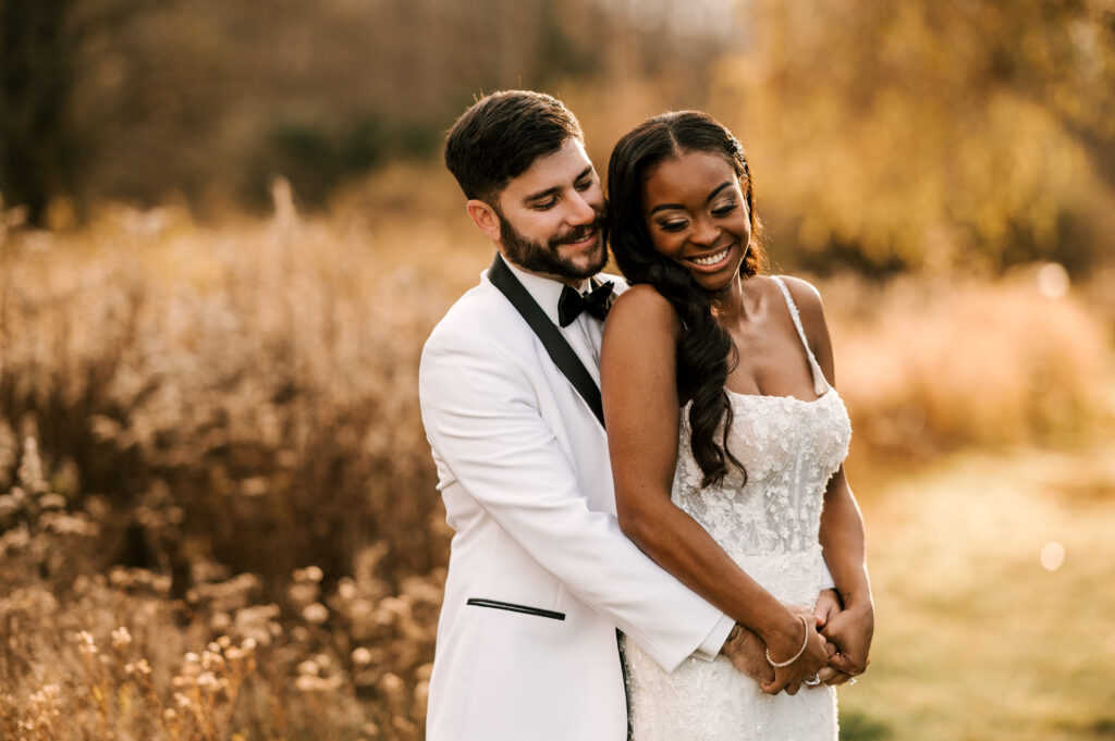 The groom hugging the bride from behind as they smile and look down.