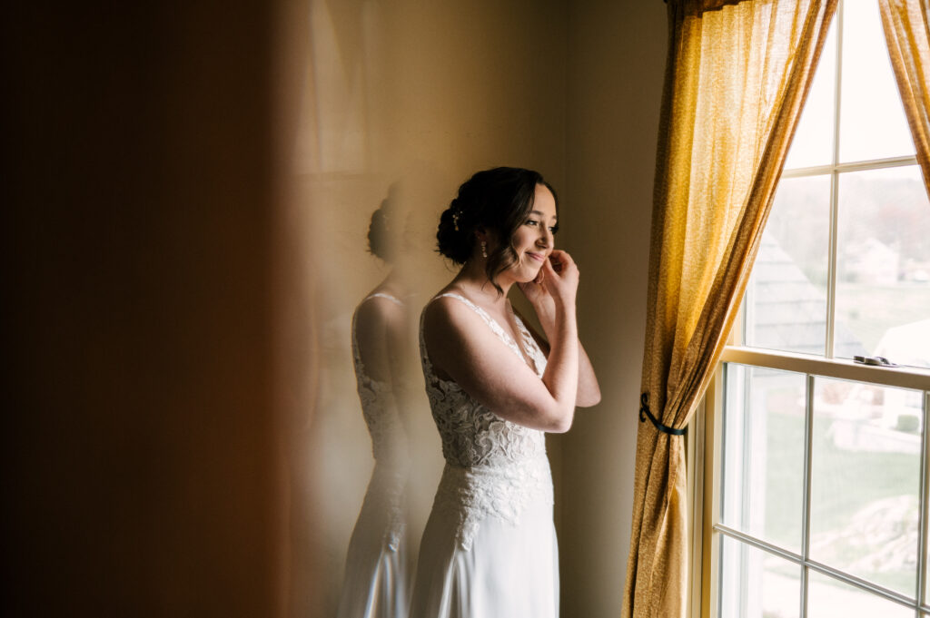 The bride smiling and looking out the window at Perona Farms Reserve as she puts her earrings on.