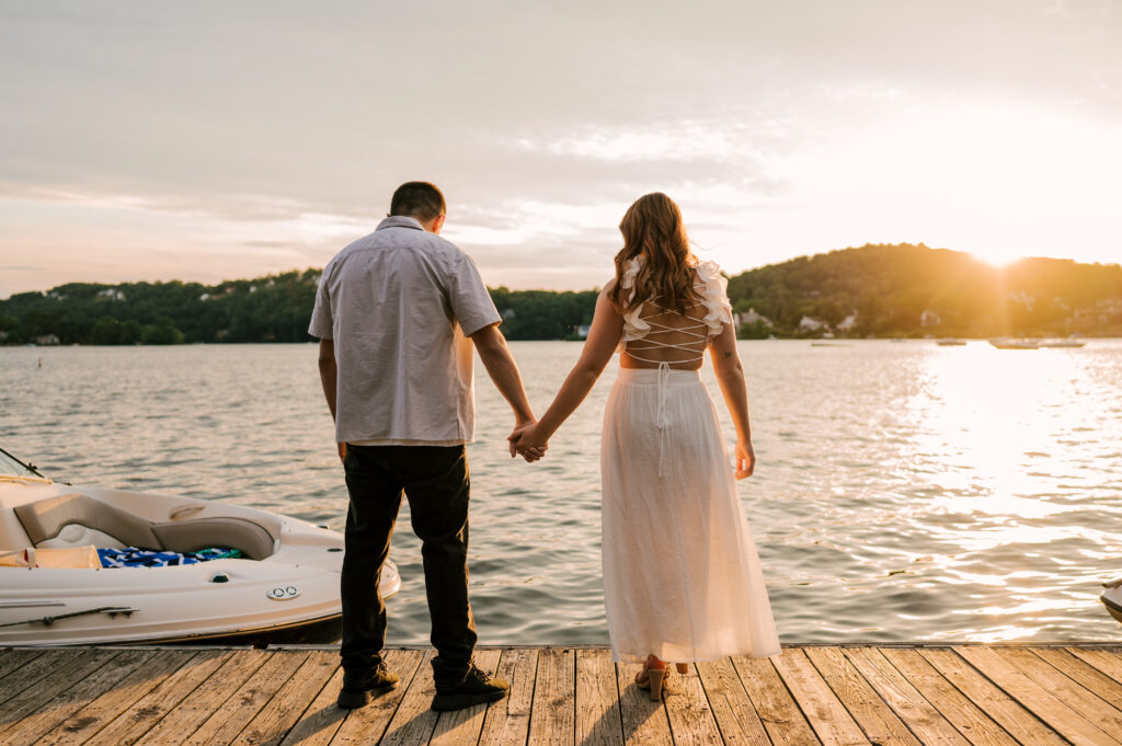 The newly-engaged couple holding hands on the dock and looking on to the water at Lake Mohawk.