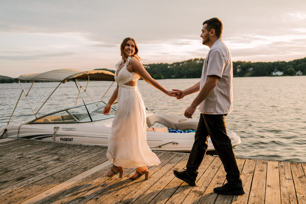 The newly-engaged couple holding hands and walking on the dock during golden-hour on Lake Mohawk.