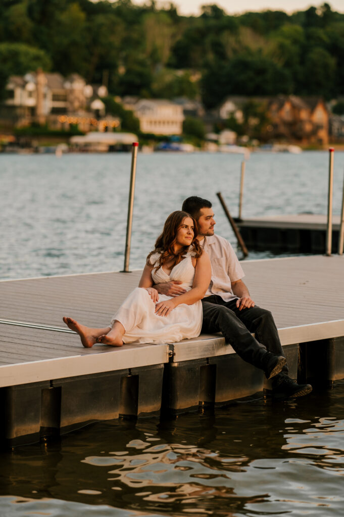The newly-engaged couple sitting on the edge of the dock and leaning on each other gazing at the sunset on Lake Mohawk.