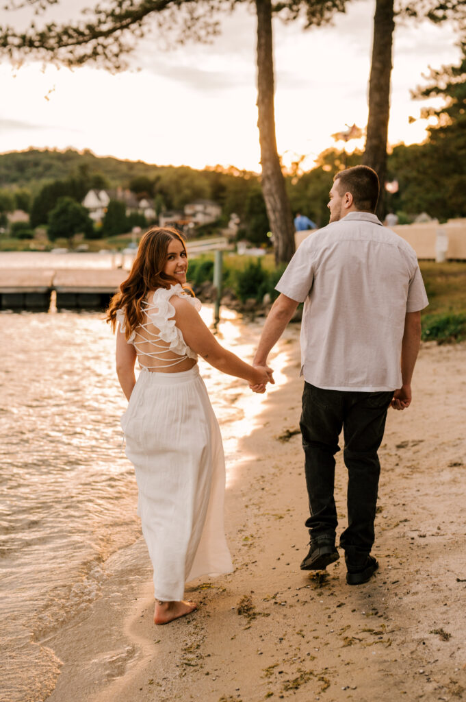 The bride-to-be looking back and holding hands with her fiancé as they walk the shore of Lake Mohawk.