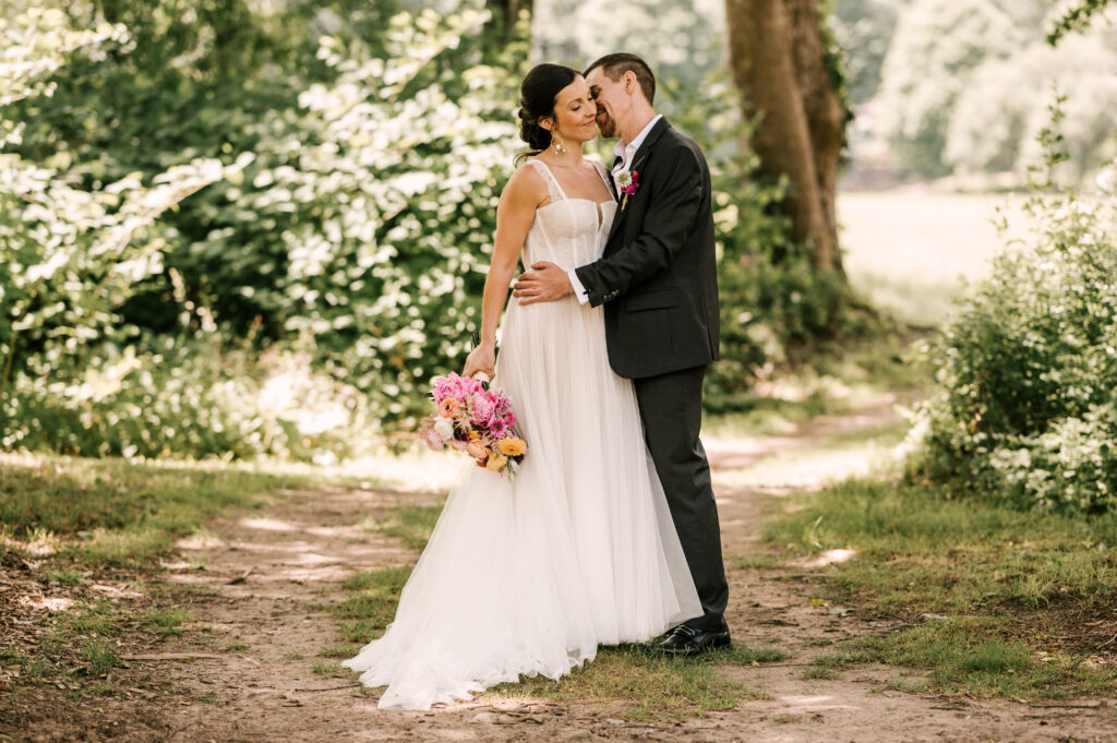 The groom kissing the bride's cheek as she smiles and holds her bouquet to the side in a woodsy area at Wallenpaupack Creek Farm.