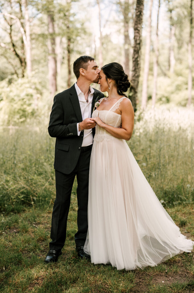 The groom kissing the bride's forehead as they close their eyes and hold hands surrounded by greenery at Wallenpaupack Creek Farm.