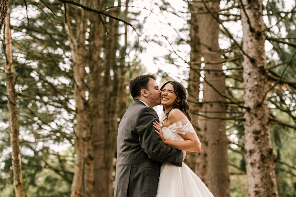 xThe groom kissing the bride's cheek as she giggles in an embrace while surrounded by trees at The Barn at Perona Farms.