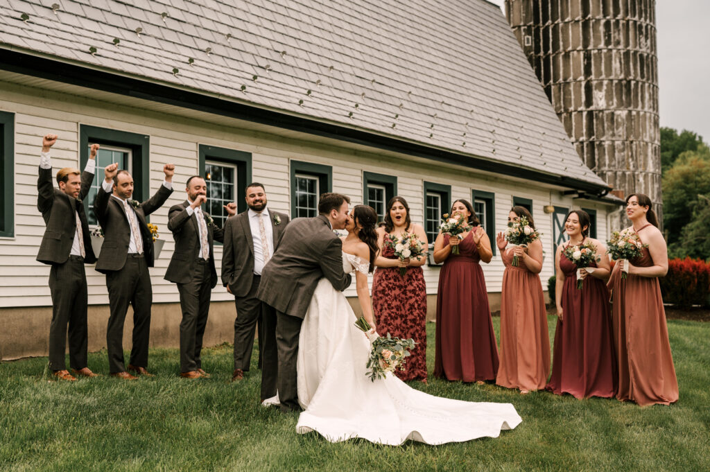 The bride and groom sharing a kiss as the bridal party cheers them on at The Barn at Perona Farms.