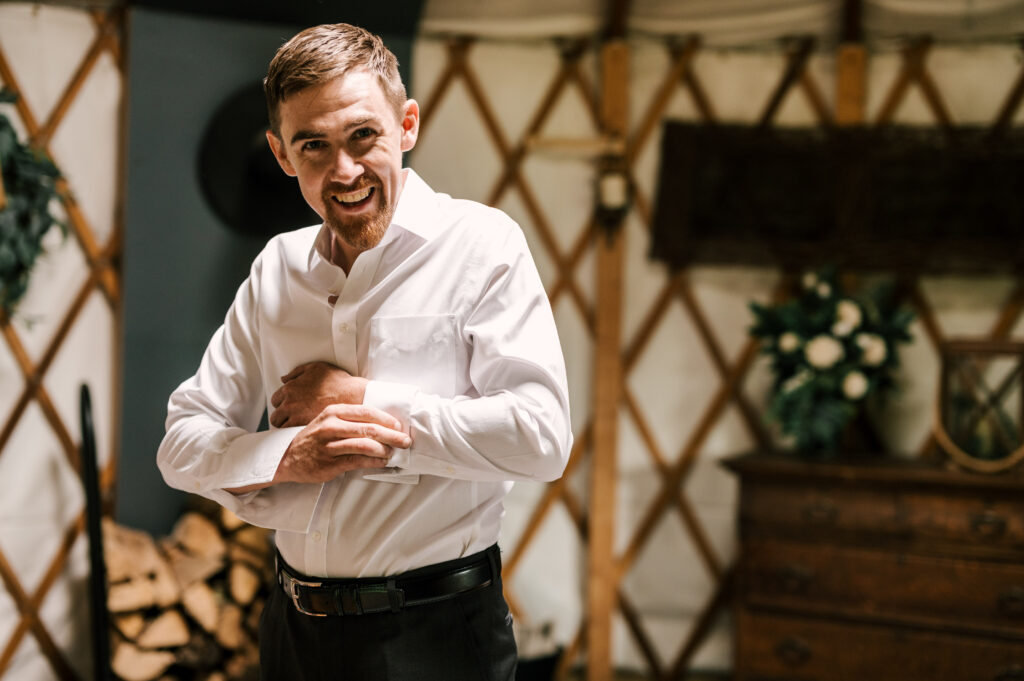 The groom smiling at the camera as he buttons his sleeves before the ceremony at Wallenpaupack Creek Farm.