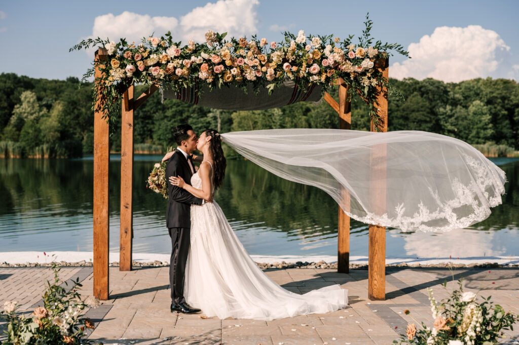 The bride and groom sharing a kiss after the ceremony by the arbor as the bride's veil flows behind her in the wind. 