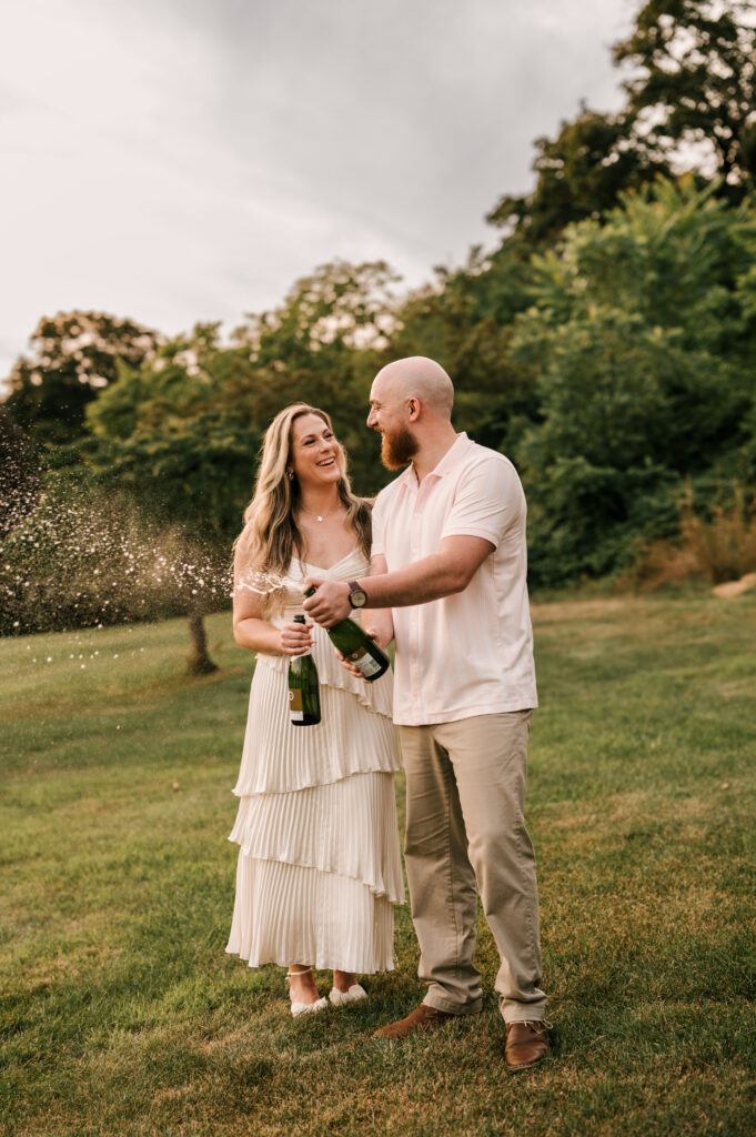The newly engaged couple looking at each other and smiling as they pop open a bottle of champagne at Skylands Manor.