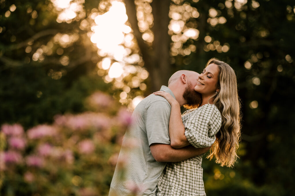 The groom-to-be kissing his fiancée as she peacefully smiles while surrounded by greenery and flowers at Skylands Manor.
