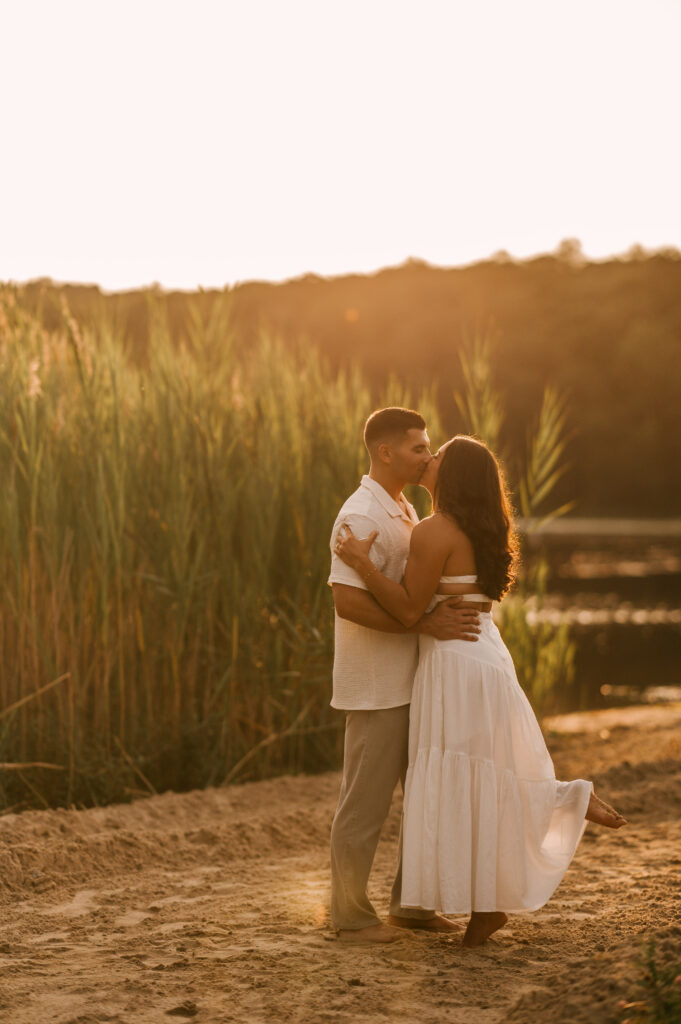 The newly engaged couple sharing a kiss as the bride-to-be kicks her foot up on the shore of Wawayanda Lake in Wawayanda State Park.