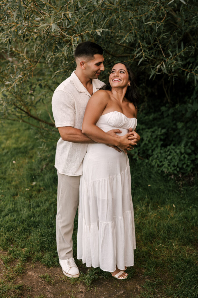 The groom-to-be hugging his fiancée from behind as they smile at each other in Wawayanda State Park.