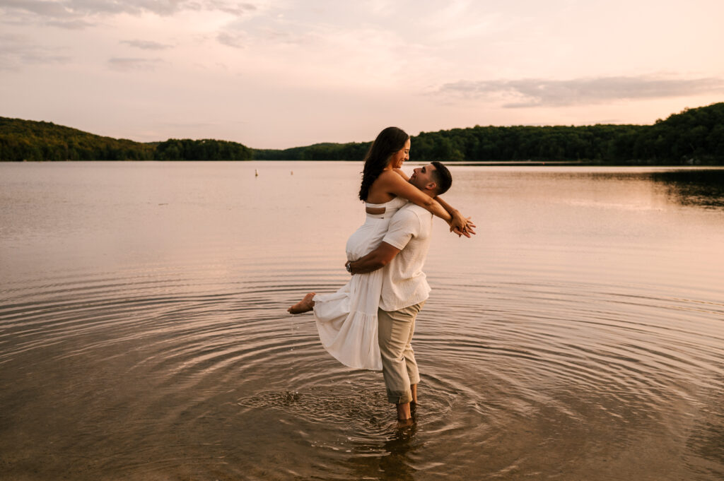 The newly engaged couple smiling and embracing on the shore of Wawayanda Lake at Wawayanda State Park.