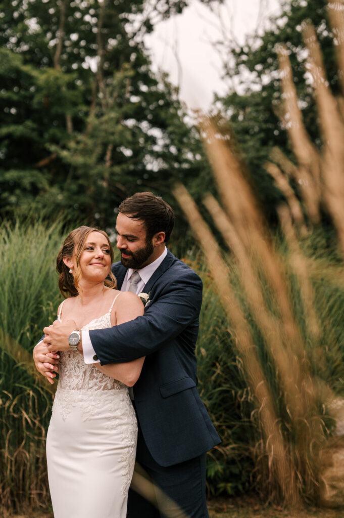 The groom hugging the bride from behind as they smile and each other surrounded by greenery at Rock Island Lake Club.