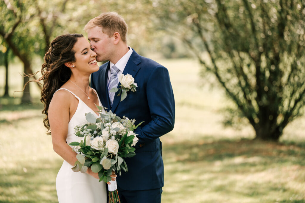 groom kissing bride during first look photos with white florals and greenery august wedding
