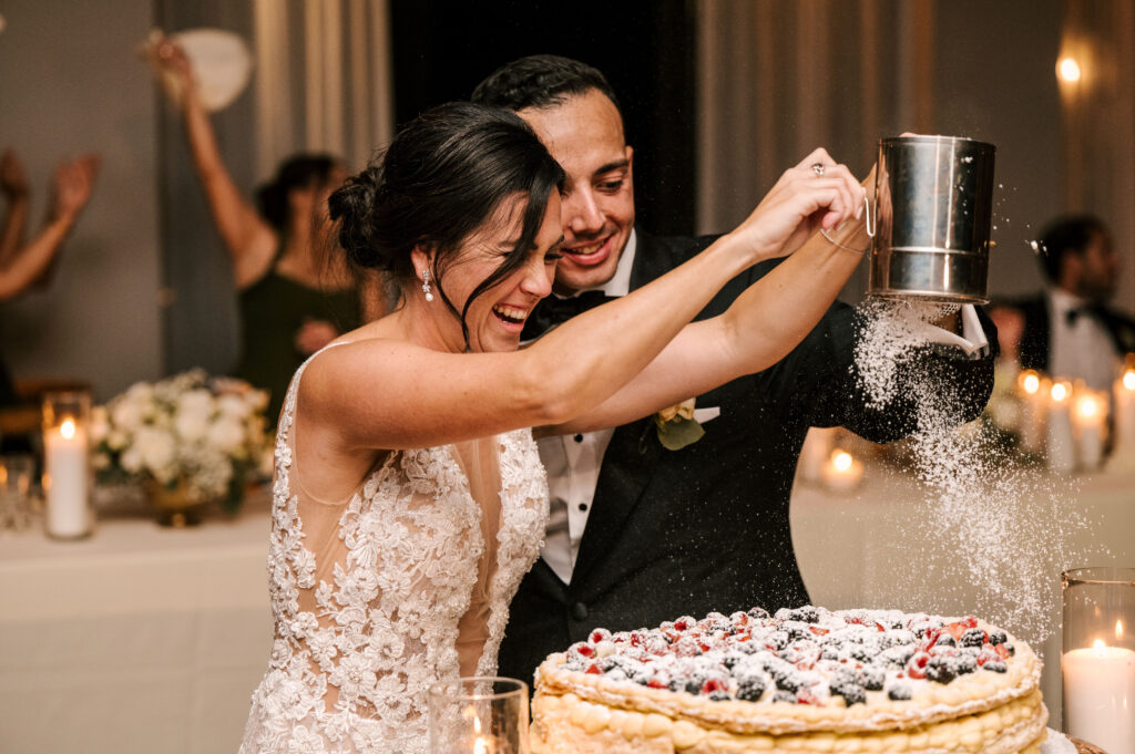 bride and groom putting powdered sugar on top of their fruit-topped wedding cake during long island wedding