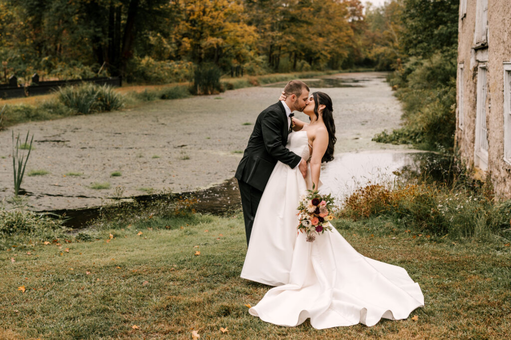 bride and groom kissing in front of picturesque river and greenery at fall nj wedding waterloo village