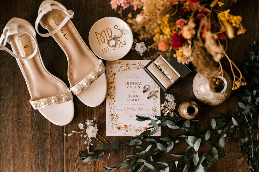 flat lay photo with bride's shoes, invitation suite, ring dish, wedding bands and florals 
