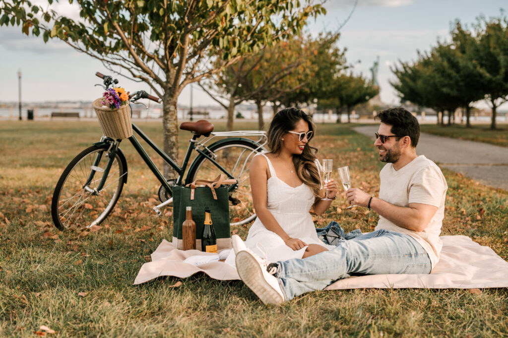 couple having a picnic on the grass with champagne glasses and trees behind them with a view of the statue of liberty - liberty state park engagement session