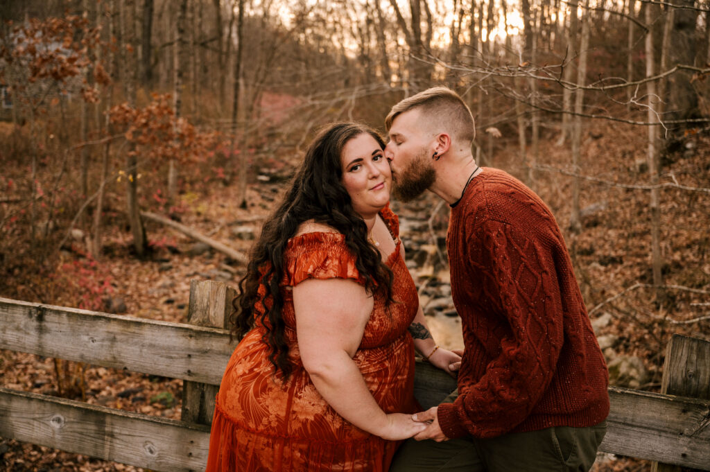 man kissing his future wife on the cheek on a bridge overlooking the fall wooded area during november engagement session in new jersey