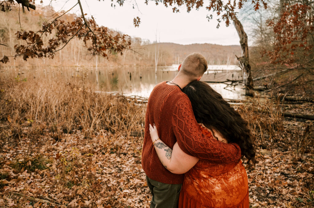 couple wearing burnt orange embracing and overlooking pond and fall leaves with backs turned at november engagement session