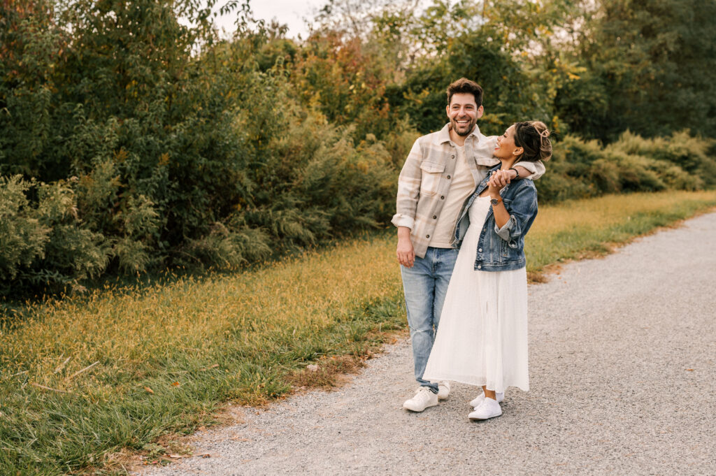 couple walking with man's arm around girl in white dress and jean jacket. greenery and fall foliage in the background. nj engagement shoot