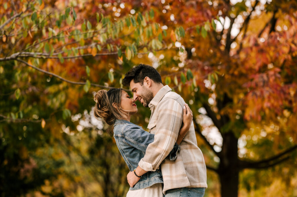 couple embracing in front of orange and red leafed trees at liberty state park during october engagement shoot