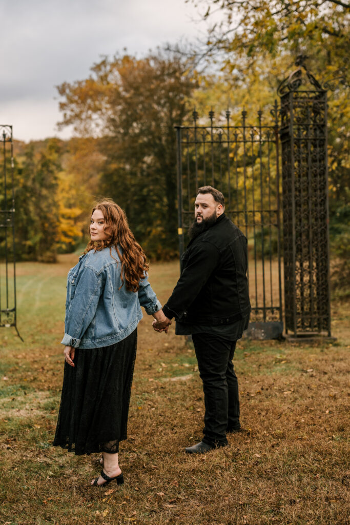 Couple stands in front of wrought iron gate at Ringwood manor for their October engagement session in New Jersey
