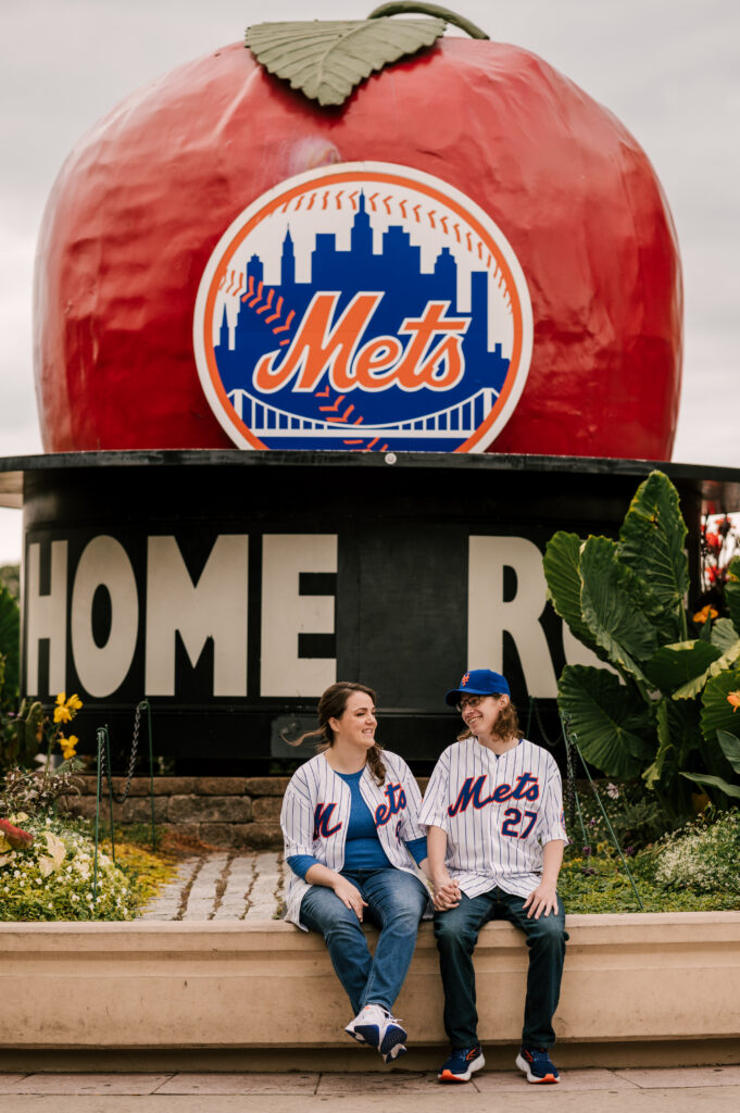 couple in Mets jerseys sitting in front of the Mets apple at Citi Field during their engagement session 