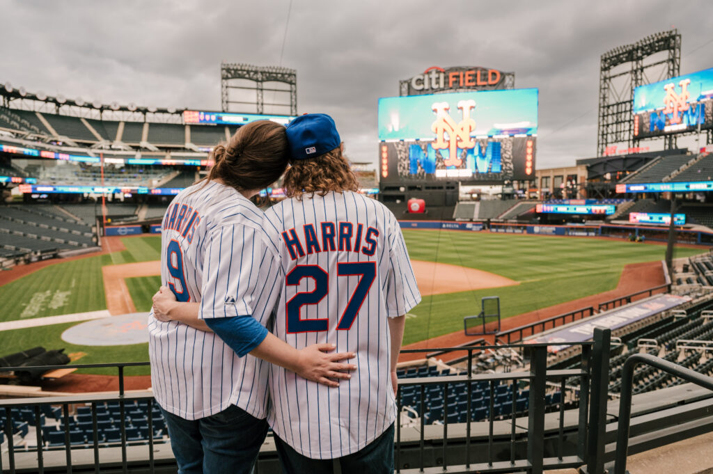 couple wearing Mets jerseys standing in the stands overlooking Citi Field during their september engagement session 