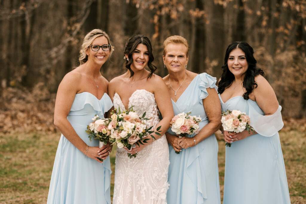 bride in strapless lace wedding gown posing with with her bridesmaids wearing light blue and holding pink & white florals