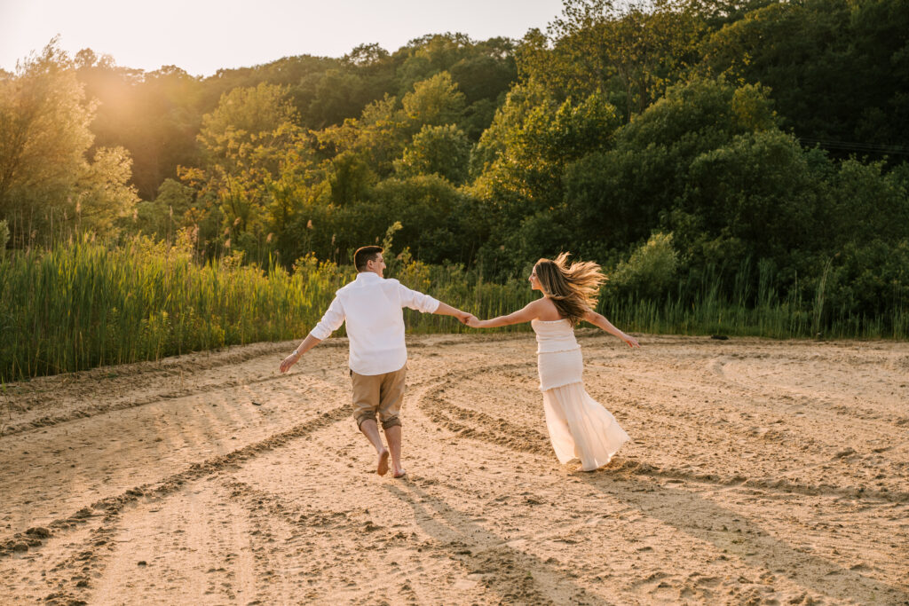 couple holding hands and running in the sand with golden hour light behind them at wawayanda state park engagement session in june