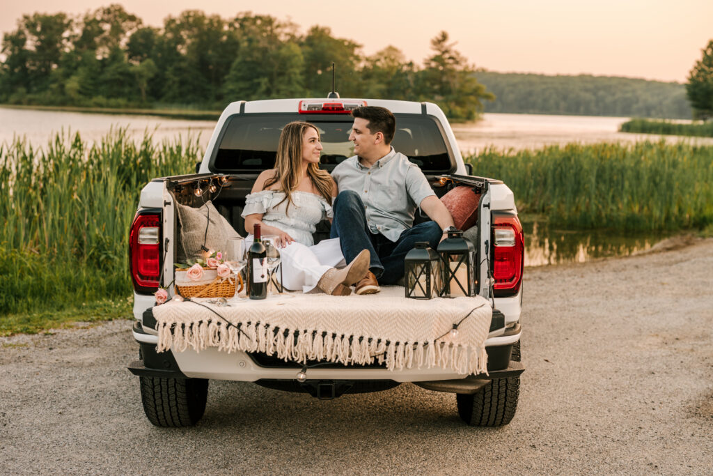 couple sitting in back of truck having a picnic with wine and pizza at their nj engagement session in the early summer